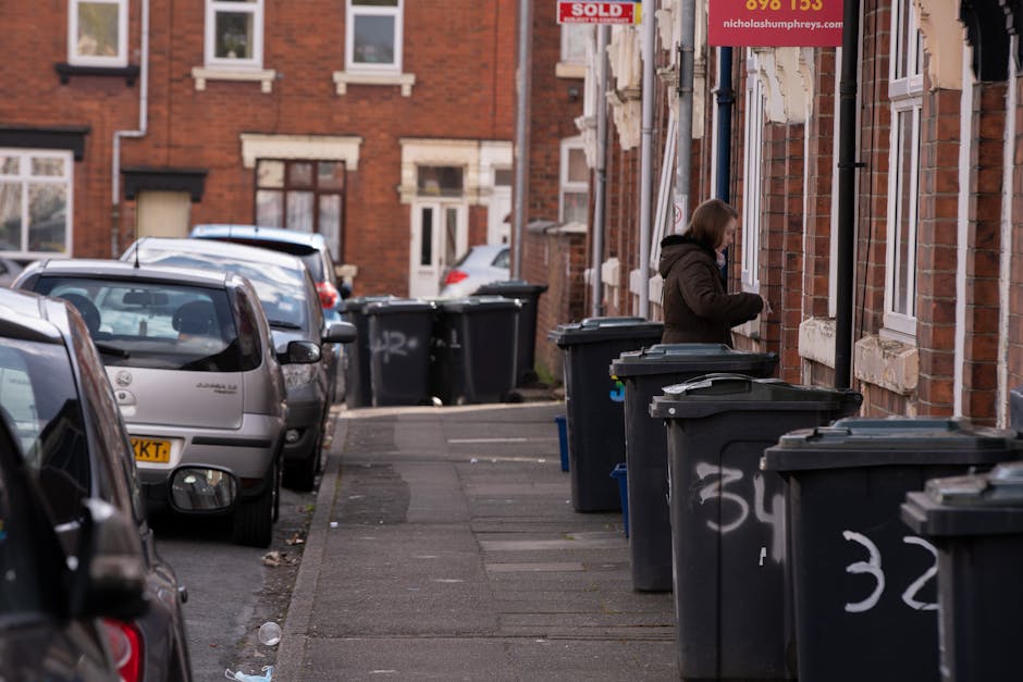 A pile of black rubbish bags and discarded debris, including a large piece of cardboard and other waste materials, situated in front of a red metal door embedded in a beige textured wall. The door dis